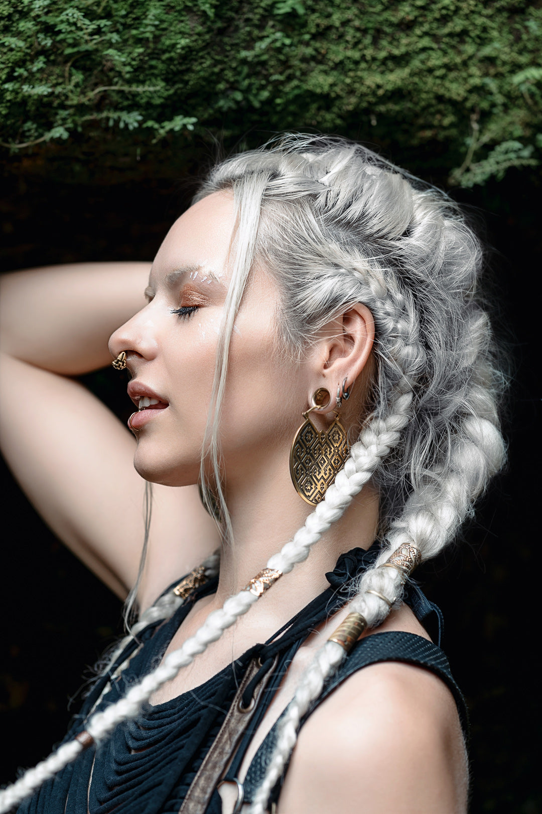 A person with long white braided hair is shown in profile with their eyes closed. They are adorned with large Kaya Movement AREVALO Shipibo Oversized Disc Hoop Earrings in Gold, a nose ring, and various beads in their hair. The background features some greenery above them.